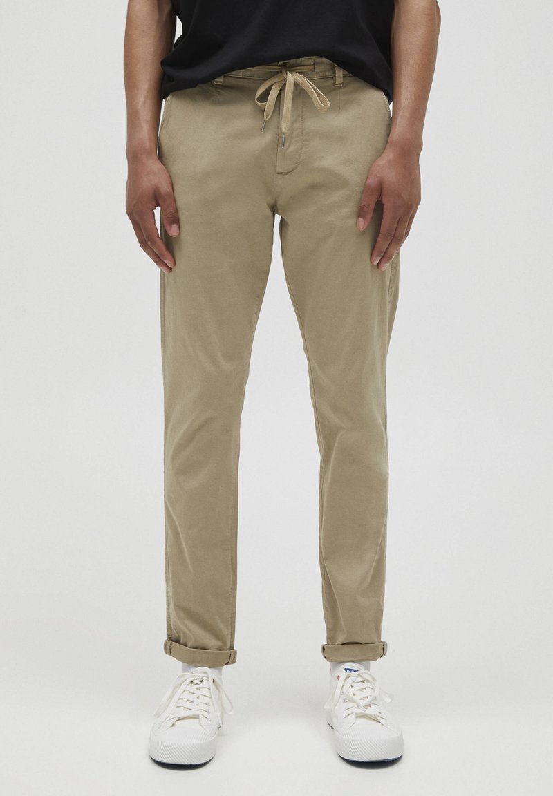 Khaki chinos med en avslappnad passform, dragsko i midjan och uppvikta manschetter. Bärs med vita sneakers. Slät bomullsdenim, minimalistisk design.