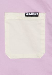 Lilla stof med en beige lomme, der har et sort mærke med teksten "CONVERSE" øverst. Lommen har afrundede kanter og synlige sømme.