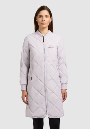 Femeie purtând o haină lungă, matlasată, de culoare gri deschis, cu fermoar frontal și buzunar la piept, stând în fața unui fundal simplu, deschis la culoare.