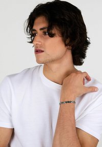 Bracciale argento con bande metalliche intrecciate, caratterizzato da un design elegante. Il modello indossa una maglietta bianca a girocollo semplice.