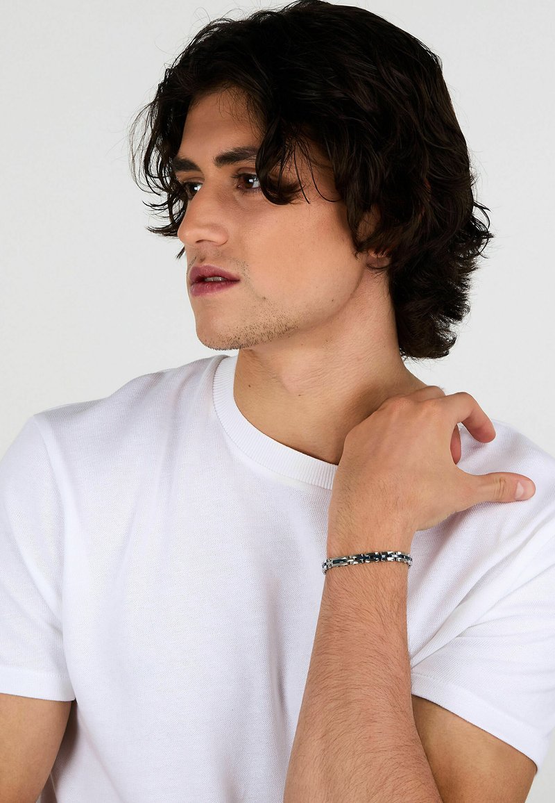 Bracciale argento con bande metalliche intrecciate, caratterizzato da un design elegante. Il modello indossa una maglietta bianca a girocollo semplice.