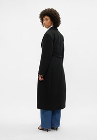 Černý dlouhý kabát s přiléhavým střihem a opaskem v pase, s čistou siluetou a kontrastní světlejší košilí pod ním, přes modré džíny.