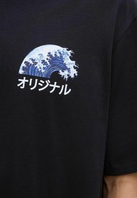 Camiseta de algodón negra con un gráfico de una ola azul y texto en japonés "オリジナル" en blanco, ubicado en el área del lado izquierdo del pecho.