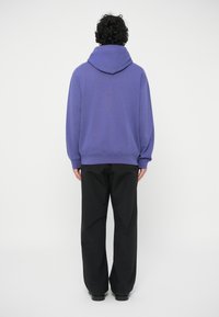 Fialová mikina s kangaroo vreckom a kapucňou, v páre s čiernymi širokými nohavicami. Hladká textura, voľný strih, minimálny dizajn.