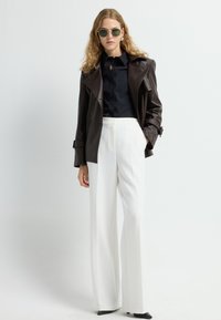 Cappotto trench in pelle marrone su una camicia nera con bottoni, abbinato a pantaloni bianchi a gamba larga e scarpe nere a punta. Occhiali da sole verdi.