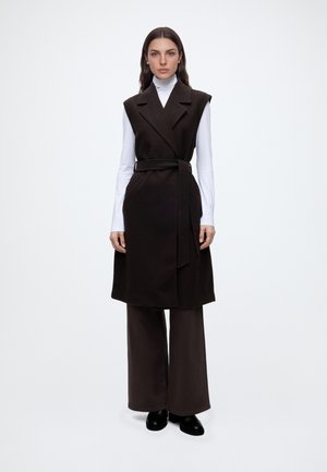 Femme portant un manteau noir sans manches ceinturé par-dessus un col roulé blanc, un pantalon noir à jambes larges et des chaussures noires, debout sur fond blanc.