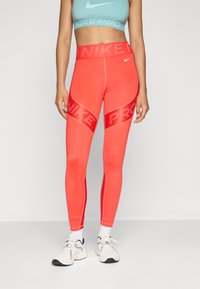 Žena u svijetlocrvenim Nike Pro tajicama, svijetloplavom Nike sportskom grudnjaku, bijelim čarapama i bijelim sportskim cipelama stoji nasuprot jednostavne pozadine.