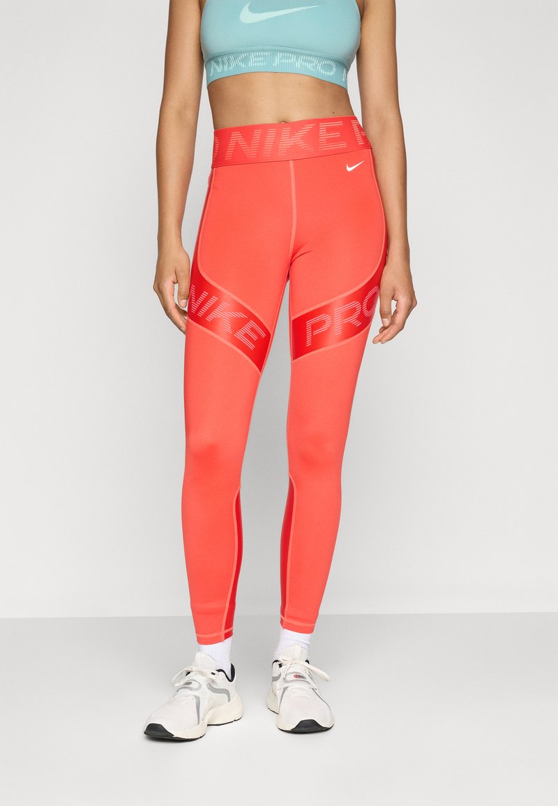 Žena u svijetlocrvenim Nike Pro tajicama, svijetloplavom Nike sportskom grudnjaku, bijelim čarapama i bijelim sportskim cipelama stoji nasuprot jednostavne pozadine.
