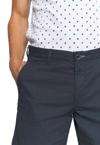 Ciemnoniebieskie spodnie o fakturowanym wykończeniu, z bocznymi kieszeniami i zapięciem na guzik, w połączeniu z białą koszulą w wzory.