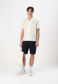 Camicia a maniche corte color crema con finitura texturizzata, chiusura a bottoni e piccolo logo giallo. Indossata con pantaloni shorts blu navy e sneakers bianche.