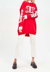 Rode oversized trui met witte opdruk; gecombineerd met witte rechte broek; zwarte platform schoenen met gouden accenten.