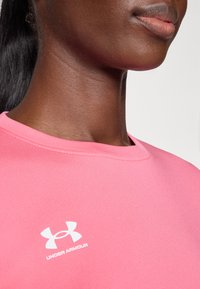 T-shirt en coton rose avec un col ras du cou et un logo Under Armour blanc sur le côté gauche de la poitrine. Texture lisse, détails de couture minimes.