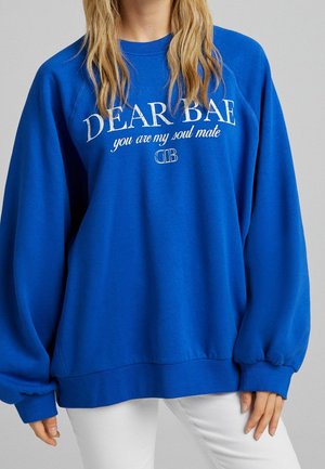 Person trägt ein leuchtend blaues Sweatshirt mit weißem Text „DEAR BAE you are my soul mate“ und weiße Hose vor einfachem Hintergrund.