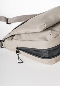 Borsa a tracolla beige realizzata in materiale durevole. Presenta una tasca con zip foderata in rete nera, una tracolla regolabile e una superficie liscia.