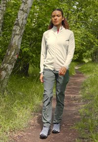 Lichtgrijs, zip-up jack met hoge kraag; olijfgroene broek; grijze wandelschoenen; wandelt op een dirt pad omringd door groen.