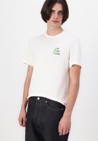 Hvid bomulds t-shirt med korte ærmer og rund halsudskæring, med et grønt grafisk print på venstre bryst, der siger "THE CLUB SCENE."