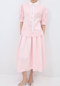 Femme portant une robe rose clair avec des manches bouffantes, un col blanc, des boutons à l'avant et des talons métalliques à bride arrière.