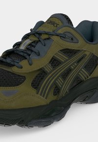 Πράσινο και σκούρο γκρι αθλητικό παπούτσι ASICS με πάνελ από δίχτυ, ριγέ σχέδια στις πλευρές και μαύρα κορδόνια σε λευκό φόντο.