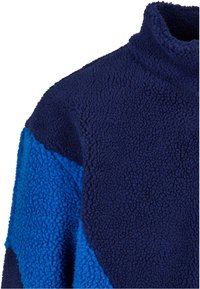Fleece pullover with high neck, featuring navy and bright blue panels, textured fabric, and a cozy, soft appearance.