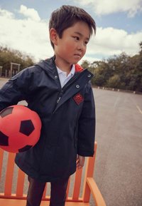 Granatowa wodoodporna kurtka z czerwonymi akcentami i logo, wykonana z lekkiego materiału, z zamkiem błyskawicznym z przodu; trzymająca czerwoną piłkę nożną z czarnymi łatami.