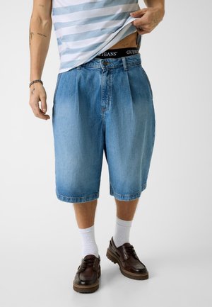 Mann trägt blaue kniehohe Jeansshorts, leicht hochgezogenen gestreiften Hemd, weiße Socken und dunkelbraune Lederschuhe.