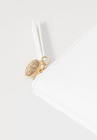 Hvit glidelåspose med teksturert overflate, med gullfarget hardware og en dekorativ charm med blomsterdesign.