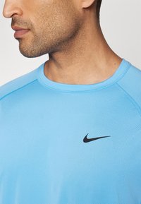 Heledsinine sportlik särk ümmarguse kaelusega, millel on must Nike logo rinnas ja tekstureeritud, hingav kangas.