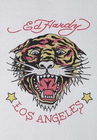 Grafika s řvoucím tygrem v černé, žluté a červené barvě s růžovými akcenty. Text pod tímto obrázkem zní "Ed Hardy" a "LOS ANGELES," se hvězdami.