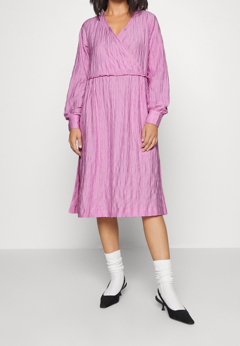 Femme portant une robe midi rose texturée à manches longues avec un col en V, associée à des chaussettes blanches et des chaussures noires à petit talon sur un fond uni.