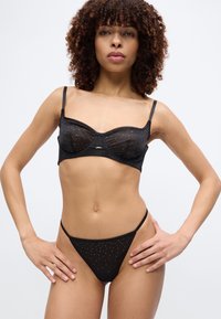 Γυναίκα με σγουρά μαλλιά που φοράει διαφανές μαύρο σουτιέν και αντίστοιχο σλιπ με ψηλό κόψιμο διακοσμημένα με μικρές λαμπερές κουκκίδες, στέκεται με τα χέρια στη μέση.