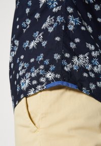 Camicia floreale blu navy con motivi botanici bianchi e blu, orlo dritto, abbinata a pantaloni giallo chiaro. Tessuto di cotone liscio.