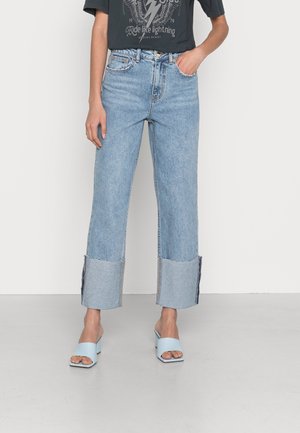 Persona que lleva vaqueros de pierna ancha de color azul claro con puños doblados, una camiseta gráfica oscura y sandalias de tacón abiertas en azul claro.