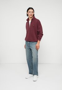 Sweatshirt bordeaux à demi-zip avec une finition texturée et un petit logo, associé à un jean en denim bleu clair et des baskets blanches.