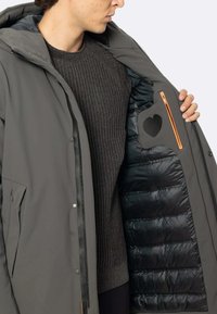 Giacca esterna grigia con una texture liscia e accessori in argento. All'interno, un rivestimento trapuntato nero e una tasca con zip arancione decorata da un dettaglio a forma di cuore.