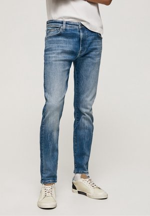 Mann in ausgebleichten, engen blauen Jeans, einem weißen T-Shirt und beigen Sneakers mit schwarzen Akzenten sowie weißen Socken.