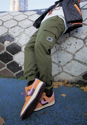 Person, der grüne Cargo-Hosen und orange-violette Sneakers trägt und an einer Wand mit sechseckigen Fliesen im Freien auf einer blauen Fläche lehnt.