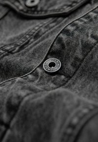Tessuto di denim nero con superficie texture, caratterizzato da un bottone metallico con la scritta "DENIM" incisa su di esso. Cuciture e pieghe visibili aumentano il dettaglio.
