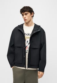 Νέος άντρας με σγουρά μαλλιά, φορώντας μαύρο μπουφάν με κουκούλα πάνω από μπεζ t-shirt με σχέδιο και χακί παντελόνι, στέκεται μπροστά σε απλό φόντο.