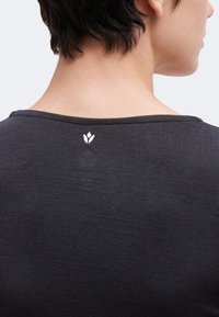 Capelli corti e scuri e collo visibili sopra una maglia nera a coste con un piccolo logo bianco ricamato al centro della scollatura posteriore.
