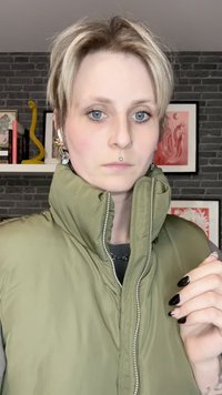 Πράσινο, επενδυμένο γιλέκο με ψηλό γιακά και ασημένιο φερμουάρ, φορεμένο πάνω από γκρι πουκάμισο. Το μοντέλο έχει κοντά, ανοιχτά μαλλιά και σκούρα νύχια.