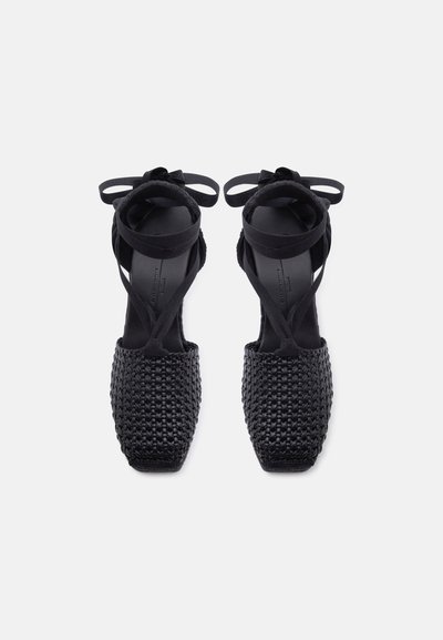 Chaussures plates noires tissées avec des bouts carrés et des lanières nouées en rubans autour de la cheville, vues de dessus sur un fond blanc uni.