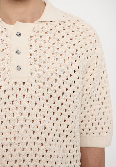 Polo en maille crème, présentant un tissage ouvert texturé, trois boutons argentés et un col côtelé. Manches courtes avec des poignets côtelés.