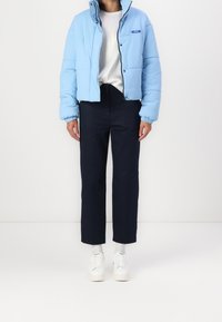 Osoba oblečená v světle modré péřové bundě, bílém tričku, černých kalhotách, bílých ponožkách a bílých teniskách stojící na bílém pozadí.