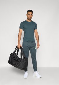 Ensemble de vêtements de sport comprenant un t-shirt ajusté en teal et un pantalon de jogging assorti avec un logo, tenant un sac de sport noir. Baskets de sport blanches.