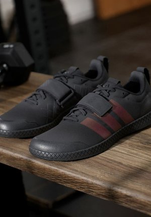 Paire de chaussures de sport noires avec des bandes rouges et une large sangle, placée sur une surface en bois à côté d’un haltère.