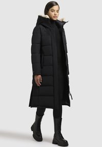 Lungo cappotto imbottito nero con cappuccio, dotato di una morbida fodera in pelliccia sul colletto, design trapuntato e zip laterali. Indossato con leggings neri e stivali.