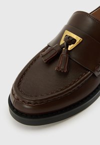 Brown leather loafer with tassel accents, gold hardware detail on the vamp, contrasting stitching, and black rubber sole.
