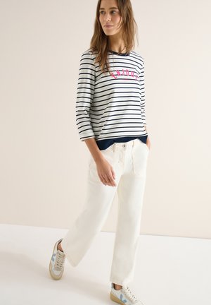 Kobieta ubrana w biało-granatową bluzkę w paski z długim rękawem z napisem "ENJOY", szerokie białe spodnie i białe trampki, spacerująca wewnątrz budynku.