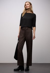 Czarna bluzka z długim rękawem zestawiona z brązowymi, wysokim stanem, kratowanymi spodniami. Modelka ma na nogach czarne botki i trzyma czarną torebkę.