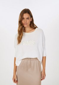 White, loose-fit top with a round neckline, featuring "PARIS" in textured fabric. Paired with a beige satin skirt. Smooth texture overall.